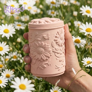 Op de afbeelding: Een roze cilindrische container met een deksel, versierd met in reliëf gemaakte zonnebloemen, bladeren en een vlinder. De container wordt in een hand gehouden, tegen een achtergrond van madeliefjes. Het woord "Nicot+" staat in de linkerbovenhoek.
