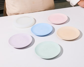 Puede incluir: Seis platos de cerámica de color pastel, incluyendo blanco, rosa claro, azul claro, lavanda, verde claro y un plato de silicona blanco.