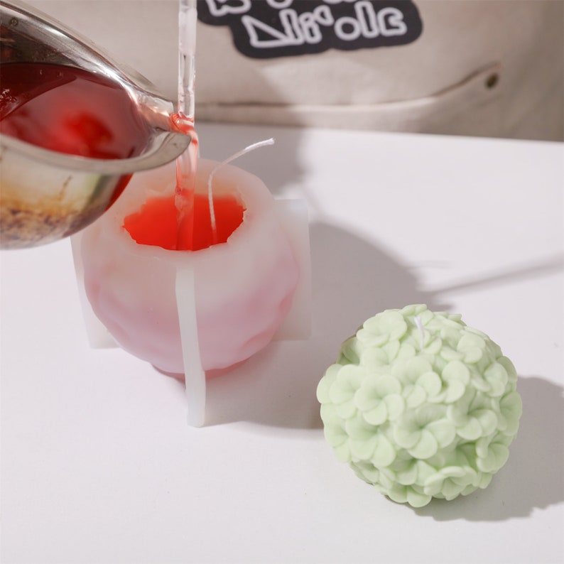 May include: A candle-making process is shown, with red wax being poured into a mold. A finished green floral candle sits nearby. The mold is a light pink color, and the finished candle is a sphere shape.