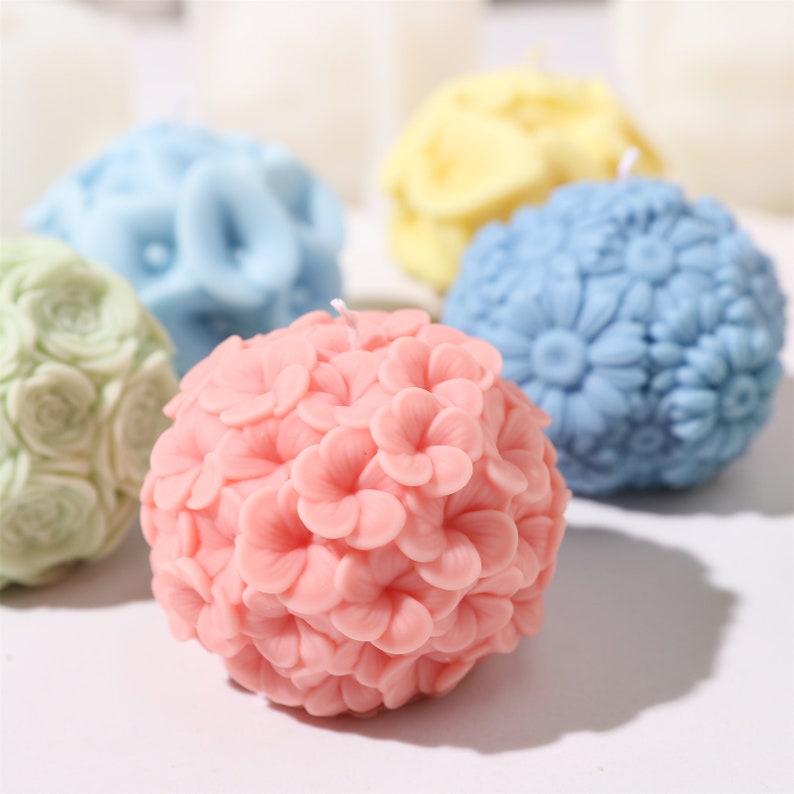 May include: A collection of spherical candles in various pastel colors. The candles are intricately designed with floral patterns, including roses, daisies, and other blossoms. The candles are pink, blue, yellow, and green, with a white wick.