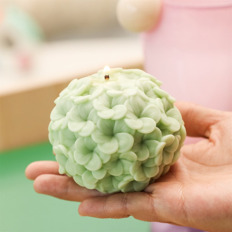 May include: A spherical, light green candle shaped like a cluster of flowers. The candle is lit, with a small flame at the top. The candle is held in a person's hand.