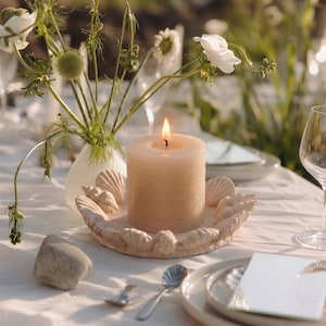 May include: A lit beige pillar candle in a seashell-shaped holder on a white tablecloth. The candle is surrounded by decorative seashells. A vase of white flowers and greenery is in the background, with a wine glass and place setting.