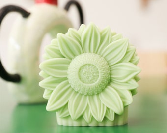May include: A light green, detailed, sculpted sunflower soap. The soap is shaped like a sunflower with a raised center and petals.