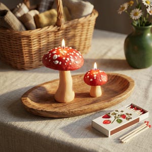 May include: Two lit mushroom-shaped candles with red caps and white spots, resting on an oval wooden tray. Beside the tray sits a rectangular matchbox decorated with a strawberry illustration and several loose wooden matches with red tips.