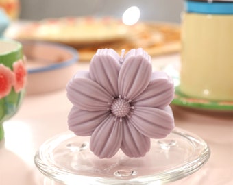 May include: A lavender-colored flower-shaped candle sits on a clear glass plate. The candle has a detailed design with petals and a center.