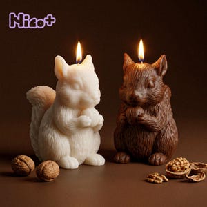 May include: Two squirrel-shaped candles, one white and one brown, each holding a nut. Both candles are lit with a visible flame. The text "Nicot" appears in the upper left corner. Several whole and cracked walnuts are placed around the candles.