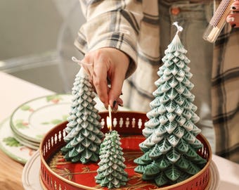 May include: Three Christmas tree-shaped candles in varying sizes, with green and white detailing, sit on a red tray. The candles are lit with a match. The tray is on a white plate, suggesting a festive holiday theme.
