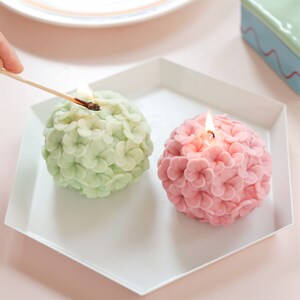 May include: Two spherical candles shaped like flowers, one light green and one pink, sit on a white hexagonal tray. A lit match is held over the green candle. The candles are unlit, with a floral design.