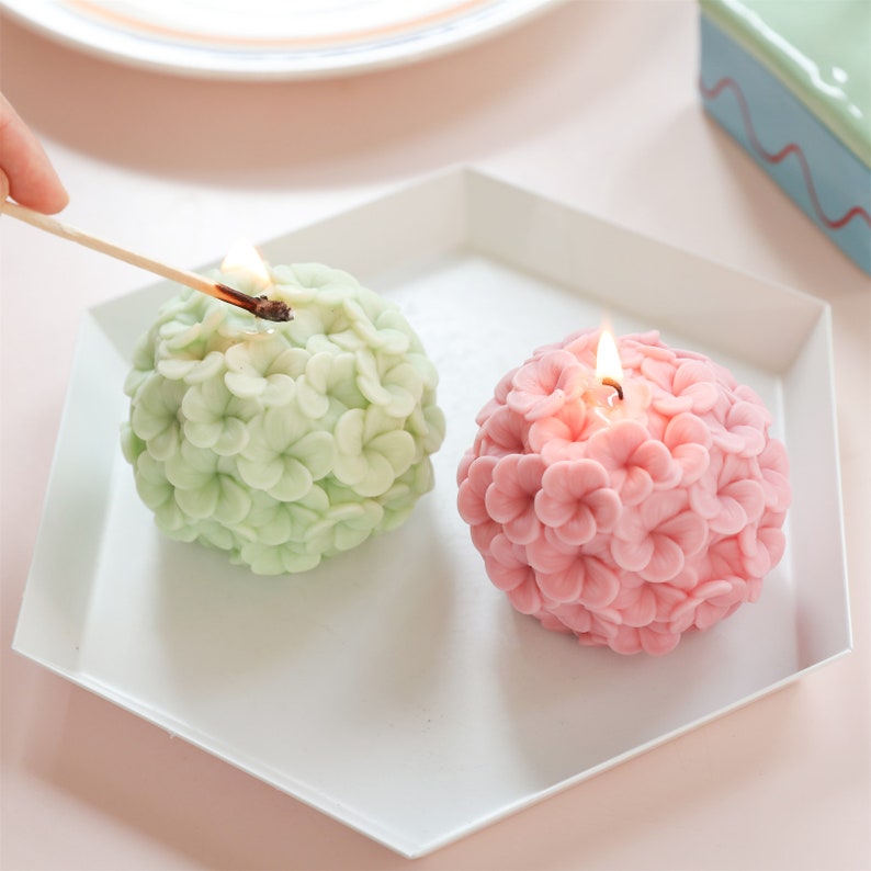 May include: Two spherical candles shaped like flowers, one light green and one pink, sit on a white hexagonal tray. A lit match is held over the green candle. The candles are unlit, with a floral design.