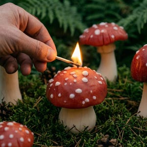 May include: A red mushroom-shaped candle with white spots being lit by a match. The candle has a thick, light-colored stem and a rounded cap. Several other similar mushroom candles are positioned nearby in a natural, mossy environment.