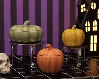 May include: Three ceramic pumpkin-shaped containers in shades of green, orange, and yellow. Each sits on a small, silver-colored table. A black house silhouette with lit windows is in the background, suggesting a Halloween theme.