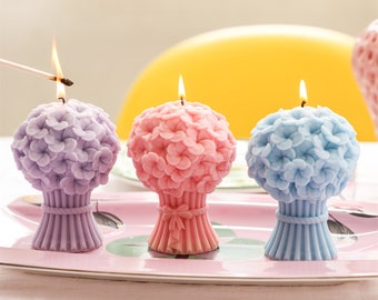 May include: Three floral bouquet candles in purple, pink, and blue. The candles are on a white plate with a pink rim.
