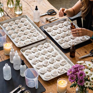 Op de afbeelding: Witte zeepjes in de vorm van schelpen liggen op metalen trays. Er zijn ook flessen, maatlepels en een maatbeker. Een persoon gebruikt een pipet om vloeistof in een fles te doen. De sc&egrave;ne speelt zich af op een houten tafel.