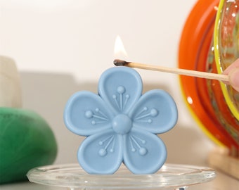 May include: A light blue flower-shaped candle is being lit with a match. The candle is sitting on a clear glass plate.