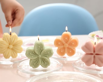 May include: Four floral-shaped candles in different colors: yellow, green, orange, and pink. The candles are on small glass plates and are lit.