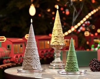 May include: Three decorative Christmas tree candles, one white, one gold, and one green, on a table with a red train in the background.