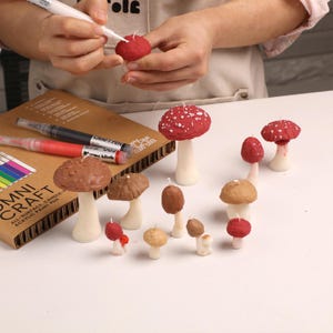 May include: A collection of mushroom-shaped candles in various sizes and shades of red and brown with white stems. An Omni Craft all-surface candle paint pen box is present, along with a white paint pen being used to add white dots to a red candle.