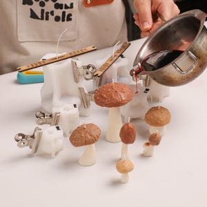 May include: A metal pouring pot dispensing dark liquid into white silicone mushroom molds. Several finished mushroom-shaped candles with brown caps and cream stems are arranged on the surface, alongside metal clips and wooden wick centering tools.