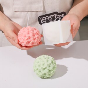 May include: A pink hydrangea-shaped candle with a white wick is held next to a white silicone mold. A green hydrangea candle sits below. The candles and mold are on a white surface. The candles are likely handmade.