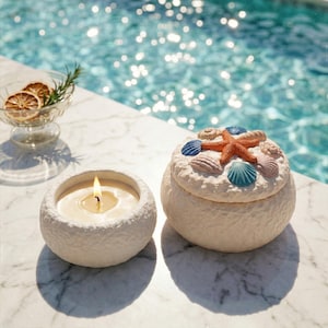 May include: A lit white candle in a textured, round holder sits next to a decorative, round box. The box is adorned with seashells and a starfish on the lid. The items are on a marble surface near a pool.