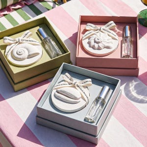 Op de afbeelding: Drie geschenkdozen met schelpvormige decoraties en glazen parfumflesjes. De dozen zijn in pastelkleuren groen, roze en blauw, elk met een wit lint. De achtergrond is een roze en witte gestreepte ondergrond.