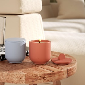 Puede incluir: Dos velas en recipientes de hormigón con tapas. Una vela es de color azul claro y la otra es de color naranja quemado. Las velas están sobre una mesa de madera.