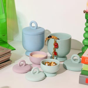 May include: A set of five pastel-coloured ceramic containers with lids and handles. The containers are in shades of blue, green, and purple. Some of the containers have a candle inside, while others are empty. There is a gold bracelet and red gemstones inside one of the containers.