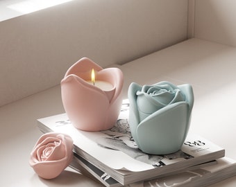Puede incluir: Dos portavelas decorativos con forma de rosas. Uno es rosa con una vela encendida en su interior y el otro es azul claro. Los portavelas están sobre una pila de revistas.