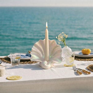May include: A pearlescent white seashell-shaped candle holder with a lit cream-colored candle. The candle holder is on a white tablecloth, with a glass of water and a lemon slice in the foreground. The background is the ocean.