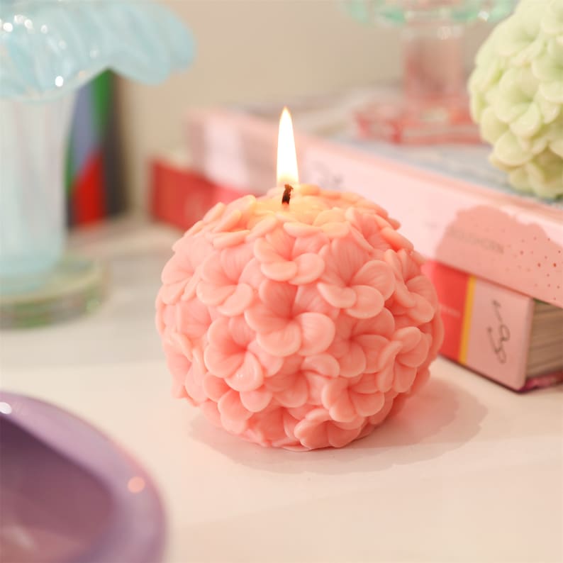 May include: A lit, spherical pink candle with a floral design. The candle is shaped like a ball covered in small, detailed flower petals. The flame is burning brightly at the top, casting a warm glow. The candle is a decorative item.