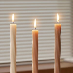 May include: Three lit, twisted taper candles in shades of beige and brown. The candles have a spiral design and are in front of a window with horizontal blinds. The flames are bright, creating a warm ambiance.