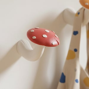 May include: A whimsical mushroom-shaped wall hook. The hook features a red cap with white spots and a curved white stem. The hook is mounted on a white wall, with a patterned fabric in the background.