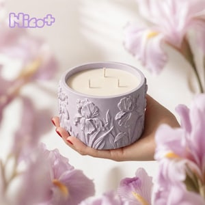 May include: A lilac-coloured candle in a round container with three wicks. The container features an iris floral design. The word "Nico+" is in the upper left corner. The candle is held by a hand, with purple flowers in the background.