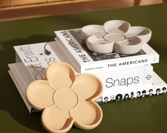 Puede incluir: Dos bandejas beige en forma de flor con seis compartimentos cada una. Las bandejas están hechas de un material liso y mate y están apiladas sobre una pila de libros. Los libros se titulan "The Americans" y "Snaps".