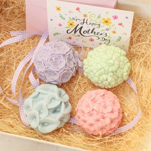 May include: A gift set for Mother's Day featuring four floral-shaped soaps in pastel colors: lavender roses, mint daisies, light blue calla lilies, and pink hydrangeas. The set includes a card that reads "Happy Mother's Day" and is nestled in a bed of straw.
