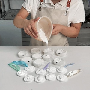 Op de afbeelding: Een persoon giet witte vloeistof in mallen. De mallen zijn gerangschikt op een wit oppervlak en hebben verschillende schelpmotieven. Er zijn ook verschillende afgewerkte witte schelpenornamenten met linten zichtbaar. Een label met "Just For You" is ook aanwezig.