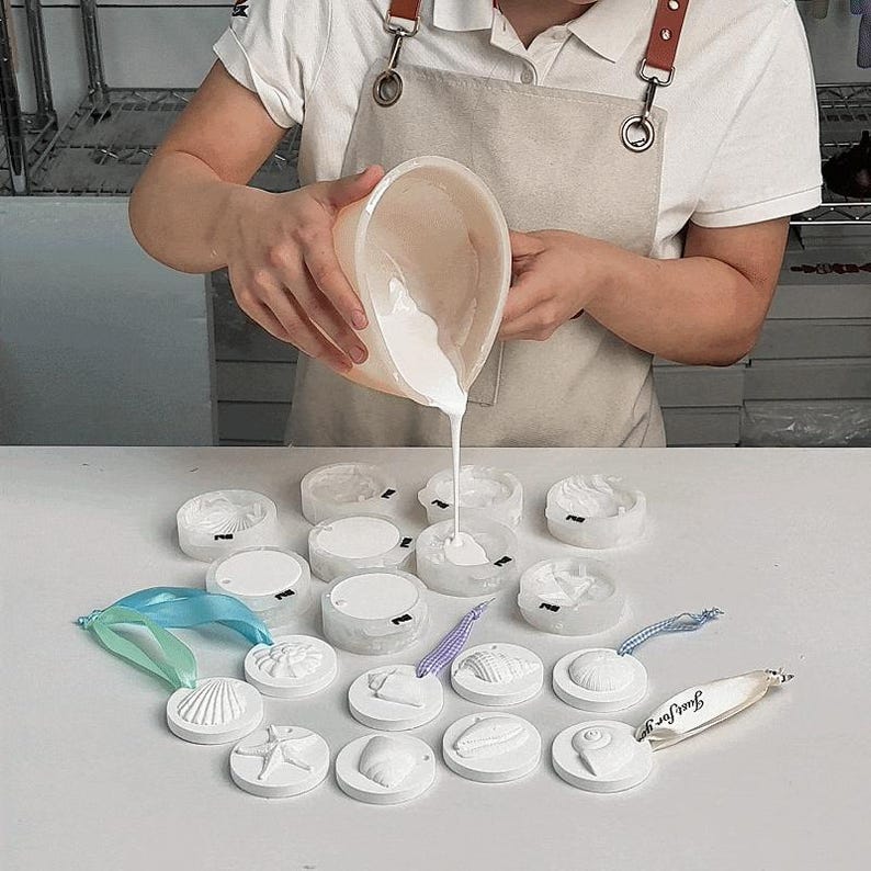 Op de afbeelding: Een persoon giet witte vloeistof in mallen. De mallen zijn gerangschikt op een wit oppervlak en hebben verschillende schelpmotieven. Er zijn ook verschillende afgewerkte witte schelpenornamenten met linten zichtbaar. Een label met "Just For You" is ook aanwezig.