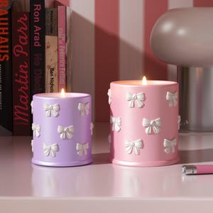 May include: Two decorative candles with a pink and purple base and white bow accents. The candles are lit and sitting on a white surface.