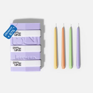 Puede incluir: Una pila de jabones de color lavanda con bandas blancas y texto en relieve, junto a cuatro velas delgadas y coloridas en amarillo, naranja, verde y morado. Una etiqueta azul indica "Eficiencia + 67%."