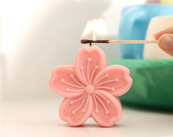 May include: A pink cherry blossom shaped candle being lit with a match. The candle has a detailed design with small dots and lines.