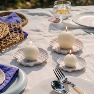 May include: Three lit, round, white candles on seashell-shaped, pale pink dishes, arranged on a white tablecloth. A fork and spoon with cream handles are in the foreground. A glass of water and a plate are also visible.