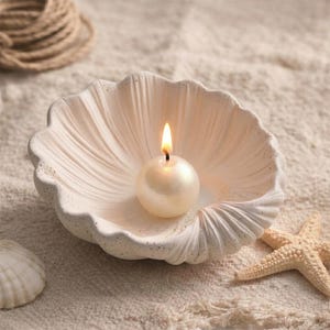 Könnte beinhalten: Ein weißer Muschelschalen-Kerzenhalter mit einer brennenden, runden, perlenartigen Kerze. Der Kerzenhalter hat eine strukturierte Oberfläche und ist von Sand und einem Seestern umgeben. Die Kerzenflamme ist hell.