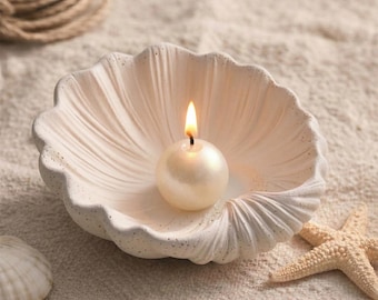 Op de afbeelding: Een witte schelpvormige kandelaar met een brandende, ronde, parelachtige kaars. De kandelaar heeft een textuur en is omgeven door zand en een zeester. De kaarsvlam is helder.