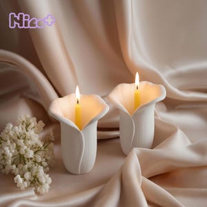 Op de afbeelding: Twee witte keramische kandelaars in de vorm van calla's, elk met een brandende gele kaars. De achtergrond is een zachte, gedrapeerde stof in een neutrale tint, met een klein boeket witte bloemen. Het woord "Nice+" staat linksboven.