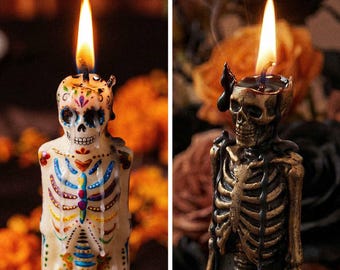 May include: Two lit candles shaped like skeletons. The left candle is cream-colored with colorful painted floral and skeletal details. The right candle is metallic bronze with black wax drips. Both candles feature a burning wick at the top of the skull.