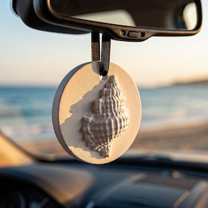 Op de afbeelding: Een ronde, beige auto-geurverfrisser met een schelpmotief, hangend aan een zwart lint. De schelp is een gedetailleerde, driedimensionale gravure. De achtergrond toont een wazig uitzicht op de zee en de lucht.