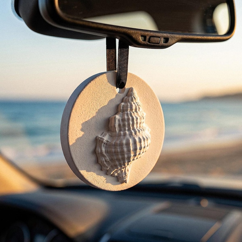 Op de afbeelding: Een ronde, beige auto-geurverfrisser met een schelpmotief, hangend aan een zwart lint. De schelp is een gedetailleerde, driedimensionale gravure. De achtergrond toont een wazig uitzicht op de zee en de lucht.