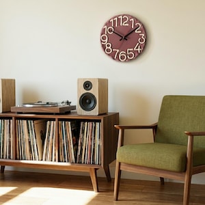 May include: A round wall clock with a dusty rose face and raised, cream-colored numbers arranged in a circular pattern. The clock features two thin black hands at the center, indicating the time against the textured, matte-finished surface.