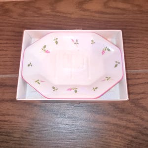 May include: A white, octagonal soap dish with a pink border and floral accents of pink roses and green leaves. The dish is presented in a white cardboard box, ideal for gifting or display.