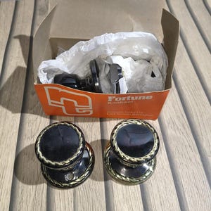May include: A set of black and gold door knobs with a circular design. The knobs have a decorative rope-like border and are displayed next to their original packaging, which features the "Fortune Knobset" logo.